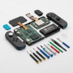 バッテリーや基板などの内部部品が並び、精密ドライバーやプラスチック製こじ開けツールなどの修理工具が周囲に配置されている