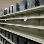CPUの箱が売れて空になった家電量販店の棚。値札に取り消し線が入った価格表示。閑散とした店内の空気感を表現